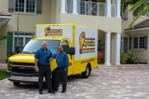 Two smiling electricians in uniform stand proudly in front of their yellow "My Electrician" work truck, parked in the driveway of a large house.