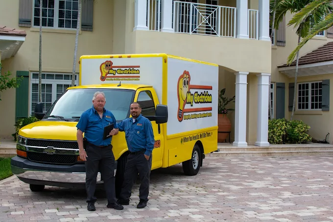 Two smiling electricians in uniform stand proudly in front of their yellow "My Electrician" work truck, parked in the driveway of a large house