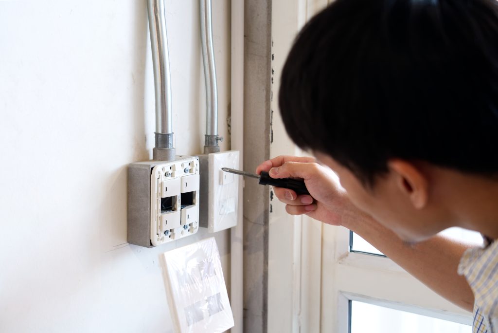 A person seen from over the shoulder uses a screwdriver to repair a wall-mounted electrical outlet with exposed wiring.