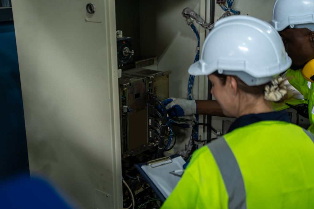 Two engineers in hard hats and safety vests inspect the open control panel of an industrial machine.