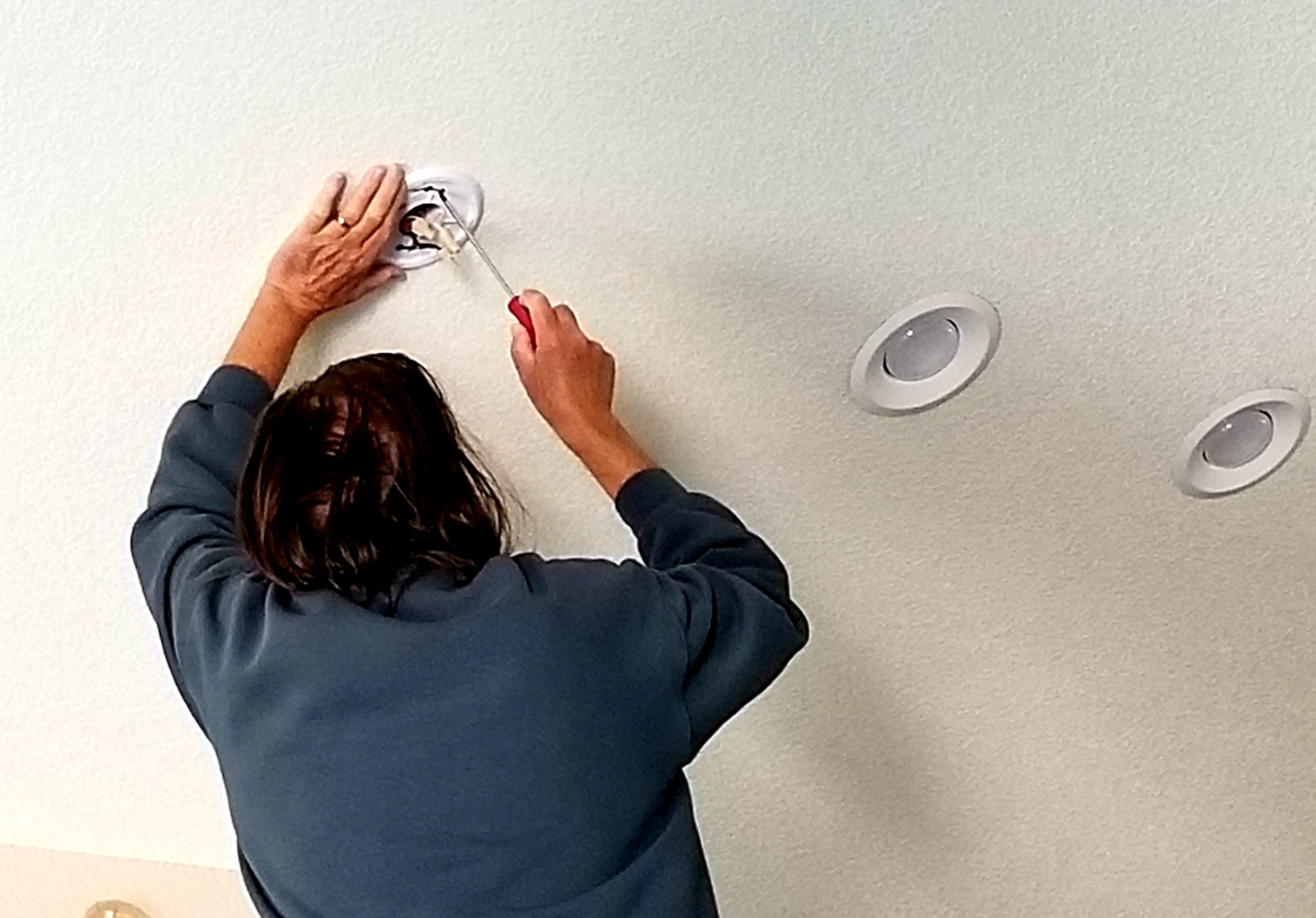 A person in a sweatshirt, seen from behind, uses a screwdriver to install a light fixture in a white ceiling.