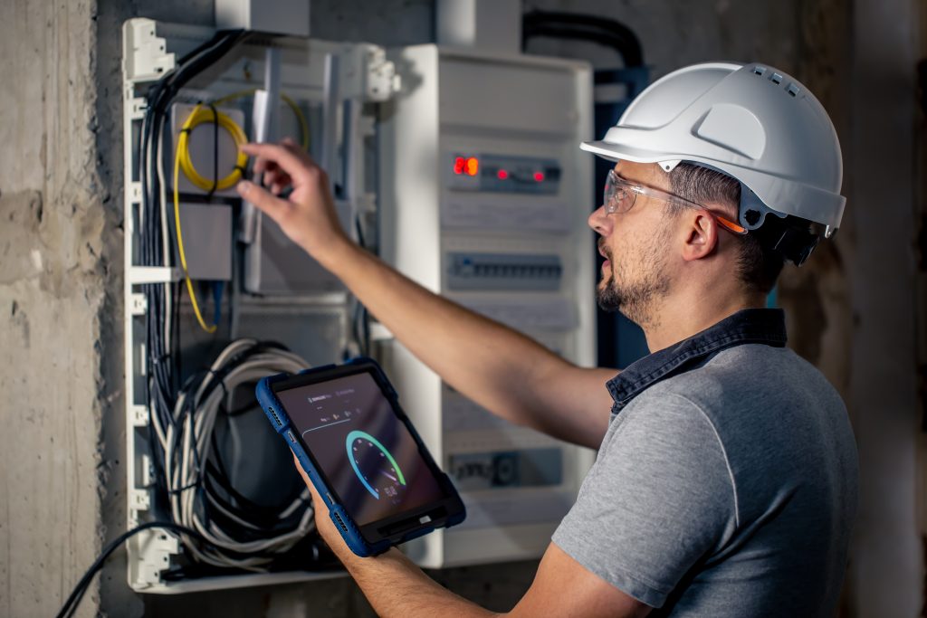 A technician in a hard hat holds a tablet while adjusting yellow wires inside an open electrical service panel.