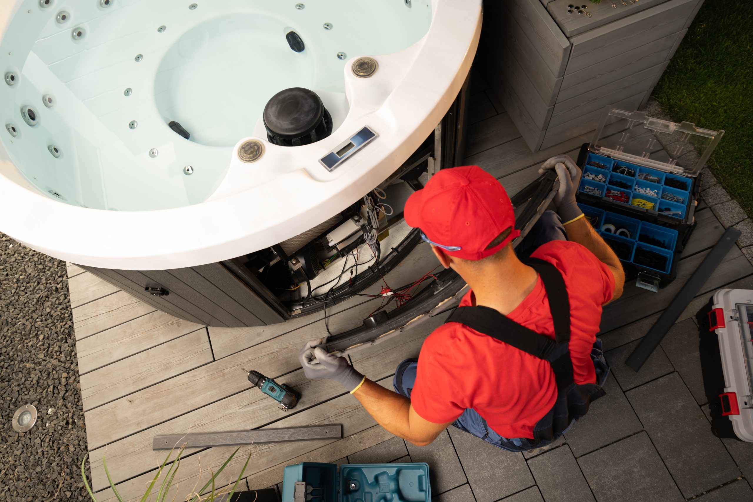 A technician seen from above works on the exposed internal machinery of a hot tub, with tools laid out on the deck beside him.