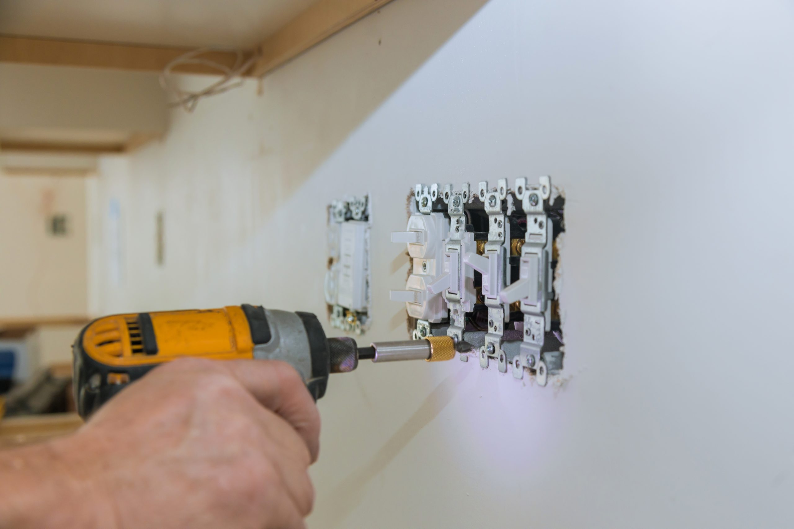 A person uses a power drill to install a bank of new light switches into an unfinished drywall wall.